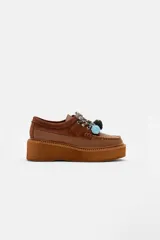 Zapato tipo mocasín con plataforma, confeccionado en cuero marrón con detalles en gamuza. Presenta cordones decorativos con nudos en colores celeste y verde oscuro.