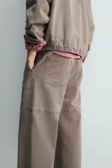 Pantalón de corte relajado en color marrón con efecto lavado. Presenta bolsillos frontales tipo cargo, cierre con cremallera y botón, y una línea horizontal de tachas metálicas a tono en la parte superior de las piernas.