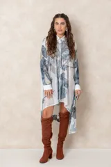 Vestido tipo túnica de corte irregular, con cierre frontal de botones y cuello camisero. Presenta un estampado botánico en tonos azules y verdes sobre fondo blanco, con detalles bordados brillantes en la parte frontal.