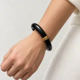 Pulsera rígida tipo brazalete de resina negra, con detalle de traba metálica dorada con diseño grabado.