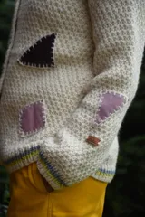 Cardigan de lana merino tejido a mano con técnica de dos agujas, de color marfil con parches geométricos de tela en tonos celestes y lila aplicados con costuras visibles. Presenta cierre frontal con botones de madera y detalles de ribetes multicolores en los bordes.
