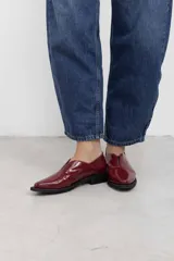 Mocasines de charol color bordó, de diseño vanguardista con corte asimétrico en el empeine, abertura en V, punta fina y suela plana con pequeño tacón.