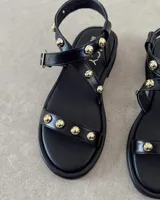 Sandalias planas de cuero negro con tiras decoradas con tachas metálicas doradas. Presentan una tira frontal sobre los dedos y una tira ajustable con hebilla alrededor del tobillo.