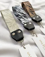 Llavero de tela con estampado animal print de cebra en tonos marrones y negros. Tiene un mosquetón metálico plateado y un aplique de cuero sintético negro con remaches plateados.