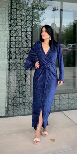 Vestido midi de fiesta en color azul, confeccionado en tela con textura satinada. Presenta un diseño cruzado con escote en V, mangas largas con puños y un detalle de fruncido en la cintura que crea una abertura frontal en la falda.