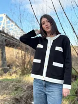 Chaqueta de punto negra con ribetes blancos, cuello redondo, cierre frontal con botones dorados, bolsillos frontales con solapa y puños acanalados.