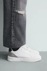 Zapatilla deportiva blanca con suela de plataforma gruesa y texturizada. Presenta detalles en gamuza gris en el lateral y un parche negro en el talón.