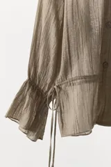 Camisa de lino color tostado con cuello subido, manga larga con puños elásticos y volantes, cintura ajustable con lazadas laterales y cierre frontal cruzado con doble botonadura.