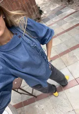 Blusa de denim liviano de corte asimétrico en tono azul, con mangas abullonadas, cuello redondo y cierre lateral con lazos para atar y botones.