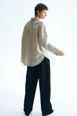 Camisa de corte oversized, color beige claro con rayas finas, cuello clásico, cierre frontal con botones, un bolsillo aplicado en el pecho, hombros caídos y mangas largas con puños abotonados.