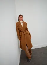 Abrigo de lana color marrón camel, de corte recto y largo midi, con solapas de muesca, bolsillos frontales con solapa y manga larga. Incluye un cinturón fino de cuero oscuro para entallar la cintura.