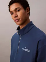 Buzo Calvin Klein de color azul marino, confeccionado en mezcla de algodón y poliéster. Cuenta con cuello alto, cierre de media cremallera y manga larga con hombros caídos. Presenta puños y dobladillo acanalados, y el logo de la marca bordado en punto de cadeneta en el pecho.