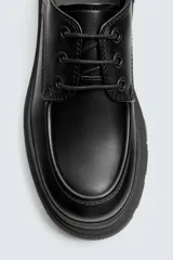 Zapato de vestir tipo derby, color negro, con detalle de bordón en la puntera. Cuenta con tirador trasero para facilitar el calce, horma redonda y suela con volumen y diseño irregular.