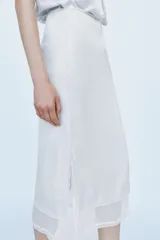 Falda midi de tiro medio, color blanco, confeccionada en hilatura de viscosa 100%, con detalle de aplicación de tejido combinado semitransparente con abertura en el bajo y cierre lateral con cremallera oculta.