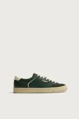 Zapatilla deportiva de estilo retro en color verde oscuro, con detalles en crema en el talón y laterales. Presenta una suela de goma en contraste de color y cordones al tono.