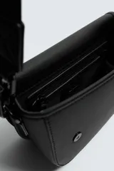 Bandolera de textura granulada color negro, con solapa frontal que incluye un cierre de cremallera. Cuenta con un asa superior fija y una correa de hombro ajustable y extraíble. El interior dispone de un bolsillo con cierre y un bolsillo plano.