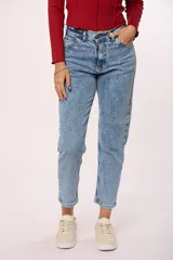 Jean de corte mom con tiro alto, confeccionado en denim de lavado claro con efecto desgastado. Presenta un diseño clásico de cinco bolsillos, cierre con cremallera y botón metálico.