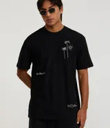 Remera negra de corte relaxed, con cuello redondo y manga corta. Presenta detalles bordados en color blanco: un diseño de cocoteros y sol en el pecho, texto en el lateral derecho y ondas en el bajo.