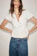 Top de color blanco con escote en pico, mangas cortas abullonadas y bajo con diseño peplum. Presenta detalle de botones dorados en la parte frontal y cierre lateral con cremallera oculta.