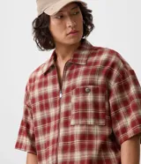 Camisa de franela de algodón con diseño a cuadros en tonos rojo y beige, de corte holgado relaxed, mangas cortas, cuello tradicional y cierre frontal con cremallera. Cuenta con un bolsillo aplicado en el pecho con detalle de botón metálico.