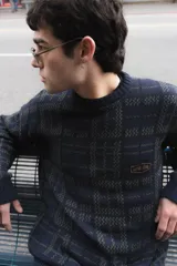 Buzo tejido de punto con diseño geométrico de cuadrillé en tonos azul y verde. Presenta cuello redondo, mangas largas y un parche de cuero sintético con logo en el pecho.