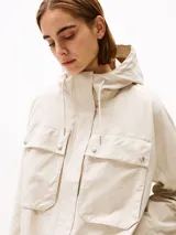 Campera beige de Tommy Hilfiger, con capucha con cordón ajustable y cierre frontal con cremallera y botones a presión. Tiene dos bolsillos delanteros tipo sobre con botón y logo metálico de Tommy Hilfiger en el puño.