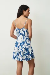 Vestido corto sin mangas, con escote en V y tirantes finos ajustables. Presenta un diseño estampado en tonos azul y blanco con motivos florales, cintura elástica fruncida y falda con vuelo y detalle de volados.
