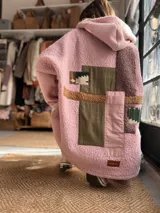 Campera con capucha confeccionada en tejido tipo corderito color rosa, con diseño de patchwork en la espalda que combina parches de tela lisa, texturizada y estampada en tonos verde oliva, marrón y rosa. Presenta un corte holgado y etiqueta de marca en el ruedo.