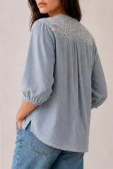 Blusa a rayas verticales en tonos rosados y blancos, con cuello redondo y cierre frontal parcial con botones. Presenta delicados detalles bordados en los hombros y mangas tres cuartos abullonadas con puños elásticos.
