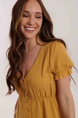 Vestido mostaza de tela texturizada, con escote en V, botones frontales y elástico en la cintura. Presenta mangas cortas abullonadas con detalle de volado en el borde.