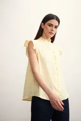 Camisa color amarillo pálido con textura arrugada, cuello mao con volados, cierre frontal con botones y mangas cortas con volados.