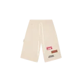 Short de felpa color arena, con cintura elástica con cordón ajustable blanco, bolsillos laterales y varios parches bordados decorativos en el frente.