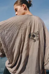 Top de viscosa color beige con escote en V, mangas murciélago abullonadas por debajo del codo y bajo asimétrico. Presenta delicados bordados con aplicaciones de abalorios y cuentas brillantes en el frente y las mangas.
