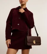 Cartera tipo shoulder bag de tamaño mediano, confeccionada en poliuretano color marrón oscuro. Presenta una solapa frontal con un detalle metálico dorado de forma orgánica y asa ajustable.