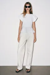 Top de popelina blanca con cuello redondo, mangas cortas abullonadas y detalle de abertura en la espalda con lazada. Presenta bajo en evasé tipo peplum y cierre lateral con cremallera oculta.