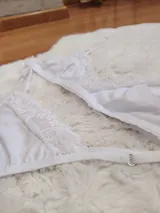 Conjunto de lencería compuesto por un bralette triangular y una tanga, ambos confeccionados en algodón blanco con detalles de encaje en los bordes. El bralette cuenta con breteles ajustables y la tanga presenta laterales regulables.