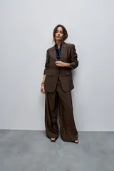 Blazer de silueta amplia y largo por debajo de la cadera, confeccionado en tela liviana con brillo sutil y caída fluida color marrón. Presenta cuello con solapas, cierre frontal, bolsillos con tapa, mangas largas y abertura posterior.