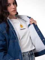 Chaqueta de jean azul con forro interior y cuello de sherpa blanco. Presenta cierre frontal con botones metálicos, bolsillos laterales y ajuste con botones en la cintura.