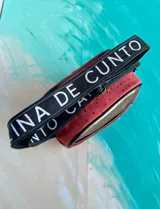 Bandolera Kaira de la línea Ottenta de Carolina De Cunto, confeccionada en cuero color rojo con diseño estilo splash en negro y bolsillo frontal superior en tono cobre. Cuenta con correa larga regulable negra con el nombre de la marca en blanco y cierre superior con cremallera.