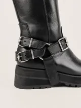 Bota de caña alta confeccionada en cuero vacuno engrasado con textura desgastada. Presenta bordados artesanales, suela con plataforma dentada y un sistema de correas con hebillas metálicas desmontables en la zona del tobillo.
