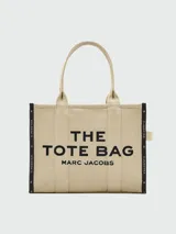 Bolso tipo tote de Marc Jacobs, modelo The Jacquard Large Tote Bag, confeccionado en tejido jacquard color tostado. Presenta el nombre de la marca y el modelo bordados en el frente en color blanco, doble asa superior y detalles de cintas laterales con inscripciones.