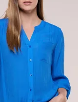 Blusa de calce relajado color salmón, confeccionada en tela liviana con textura sutil. Presenta escote en V con abotonadura frontal, bolsillo parche en el pecho, mangas largas regulables con presilla y ruedo con opción de anudar en el frente.