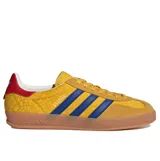 Championes Adidas Gazelle Indoor de diseño clásico, con exterior en tono amarillo con estampado geométrico sutil, tres bandas laterales en azul y detalle en rojo en el talón. Presenta puntera de gamuza y suela de caucho color caramelo.