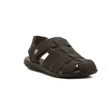 Sandalia tipo pescador para hombre, color marrón oscuro, con tiras entrelazadas en la parte superior y cierre de velcro ajustable en el lateral. Posee una suela gruesa de goma en tonos marrones.
