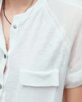 Remera blanca de lino con cuello mao, media cartera con broches metálicos y manga japonesa. Presenta un bolsillo frontal tipo plaqué, recortes laterales en sisa con detalles de encaje y ruedo redondeado con corte escalonado.