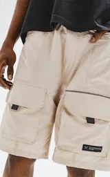 Bermuda color beige de calce relax-fit, con cintura elástica y cordón de ajuste negro. Presenta grandes bolsillos cargo con fuelle en las piernas y un cinturón de cinta negra con hebilla de plástico.