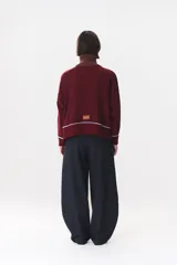 Buzo de tejido de punto en color azul marino, con cuello alto y cierre frontal parcial. Presenta un diseño con franjas en color marrón y detalles de líneas en rojo en el pecho, hombros y ruedo. Tiene corte holgado y mangas largas con puños acanalados.