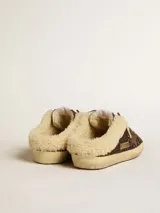 Zapatilla tipo sabot de estilo urbano, color marrón oscuro con acabado de glitter. Presenta la estrella lateral en gamuza marrón y forro interior de shearling color crema. La suela es de goma envejecida.