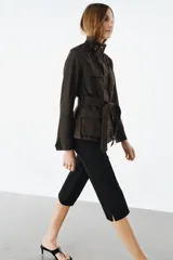 Chaqueta de estilo safari confeccionada en mezcla de algodón con acabado encerado. Presenta cuello alto con trabilla ajustable y hebilla metálica, cierre frontal con cremallera y botones ocultos bajo solapa, bolsillos de plastrón con solapa en el delantero y cinturón ajustable del mismo tejido para anudar en la cintura.