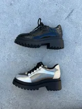 Zapato mocasín plateado con cordones negros, plataforma track negra y detalles en cuero color crema.